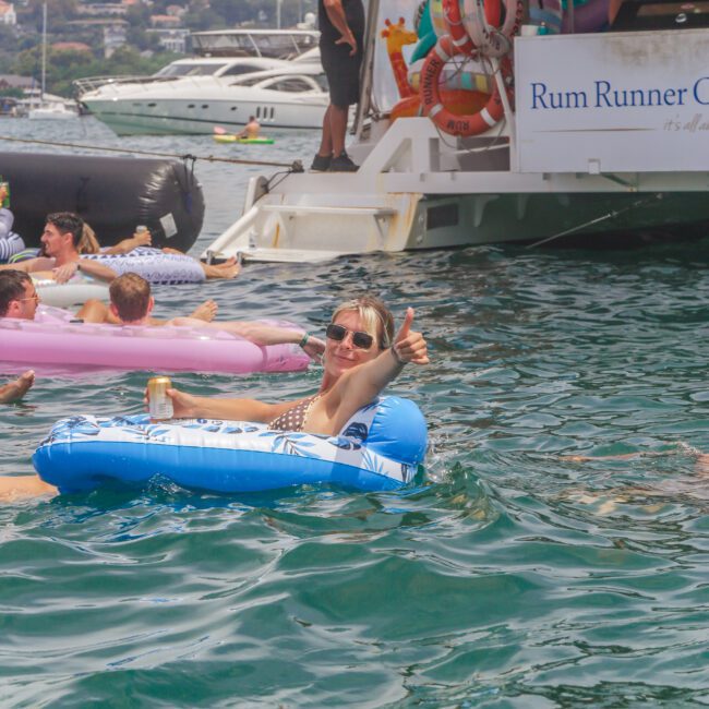 A woman floats on a blue inflatable ring in the water, smiling and giving a thumbs-up. Several people are on inflatables nearby, and a boat labeled "Rum Runner" is anchored in the background.