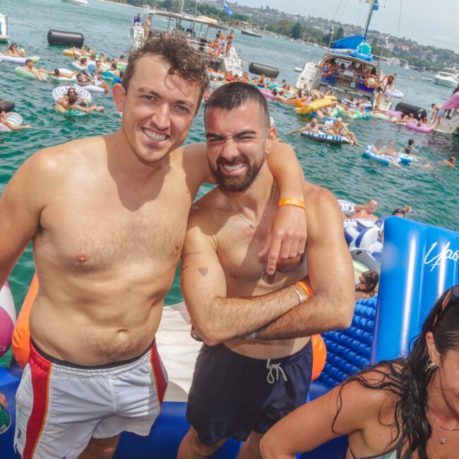 Two smiling men in swim trunks pose together on a floating platform at a lively pool party, surrounded by people swimming and relaxing on inflatables. Boats and swimmers are visible in the sunny background.