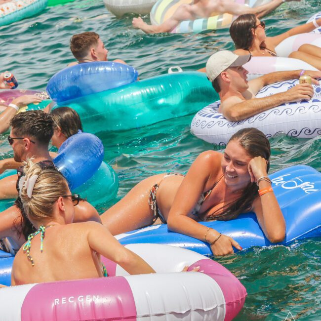 People relaxing on colorful inflatable tubes and floats in a crowded outdoor pool or body of water, enjoying the sunny weather. Most are wearing swimsuits and sunglasses, with a lively, social atmosphere.