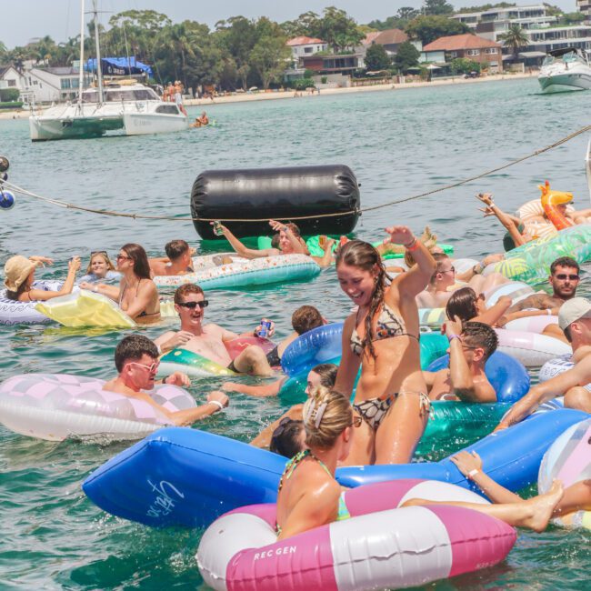 A group of people relax and socialize on colorful inflatable pool floats in the water near the shore on a sunny day, with boats and houses visible in the background.