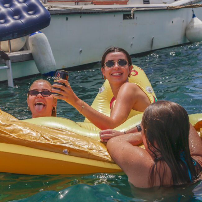 Three women relax on a yellow inflatable float in the water near a boat. One woman smiles and holds up a drink, another sticks out her tongue, and the third leans on the float. Other people are visible on a boat in the background.