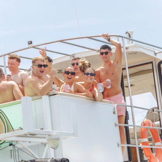 A group of smiling, swimsuit-clad people enjoying drinks and posing on the deck of a boat under a sunny sky, with pool floats and a lifebuoy visible nearby.
