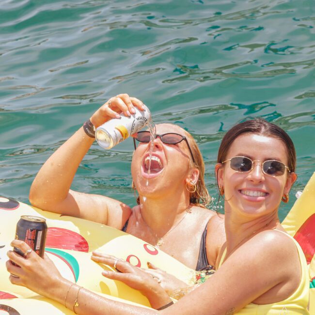 Two women in sunglasses relax on a pizza-shaped pool float in the water. One woman pours a drink into her mouth from a can, while the other smiles happily at the camera, holding a can in her hand.