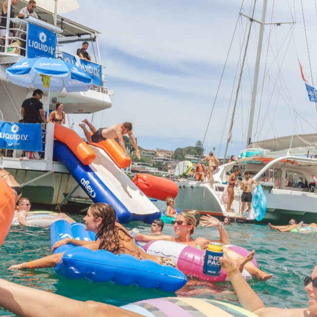 People enjoy a lively yacht party, swimming and lounging on colorful inflatables in the water, with others sliding off a boat’s slide. Boats, banners, and blue skies complete the festive summer scene.