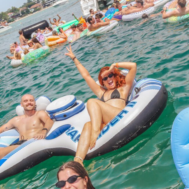 People relaxing on inflatable floats in a lively lake scene; a woman in a black bikini smiles and flashes a peace sign, while a man lounges beside her. Many others float and mingle in the background on a sunny day.