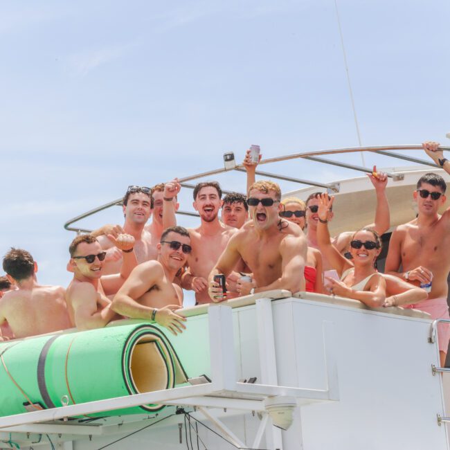 A group of people in swimsuits are smiling, cheering, and holding drinks while gathered on the deck of a boat under a sunny sky, with rolled-up green mats visible in the foreground.