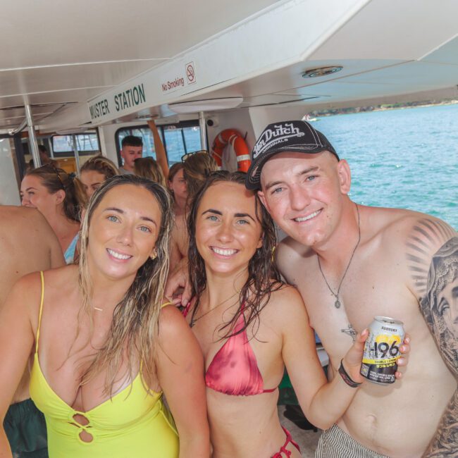 A group of people in swimwear smile and pose together on a boat, with water visible outside. One man holds a drink can and has a tattooed arm. The atmosphere is lively and casual, suggesting a fun, social outing.