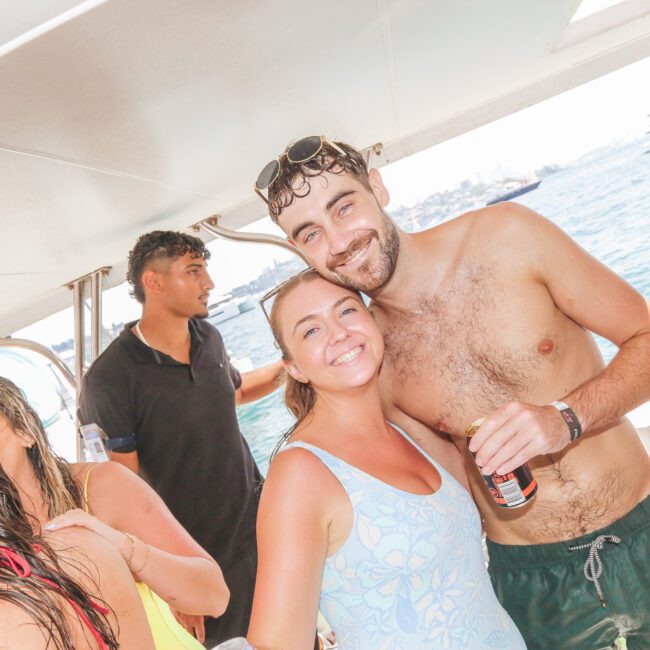A smiling man and woman in swimwear pose together on a boat with others around them. The man holds a drink and wears sunglasses on his head. The sea and other boats are visible in the background.