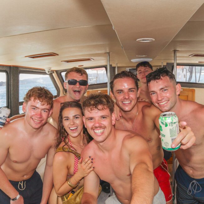 A group of smiling young adults in swimsuits pose for a selfie inside a boat. They are holding drinks and appear to be enjoying a sunny day on the water, with ocean and boats visible through the windows behind them.