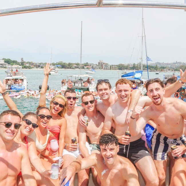 A group of young adults in swimwear smiling, holding drinks, and posing together on a boat. More boats and people swimming are visible in the sunny background on the water. Everyone appears to be celebrating and having fun.
