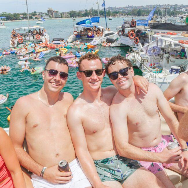 Three smiling men in swim trunks and sunglasses sit together on a boat, holding drinks. Behind them, people lounge on inflatables and swim in a busy, sunny harbor filled with boats.