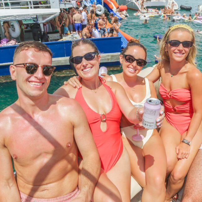 Four people in swimsuits and sunglasses sit and smile on a boat, holding drinks. Behind them, others swim and relax near anchored boats in clear, blue-green water on a sunny day.