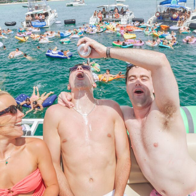 Three people enjoy a lively boat party; one man pours a drink into another’s mouth while a woman in sunglasses laughs. In the background, boats and many people float and socialize on the water.