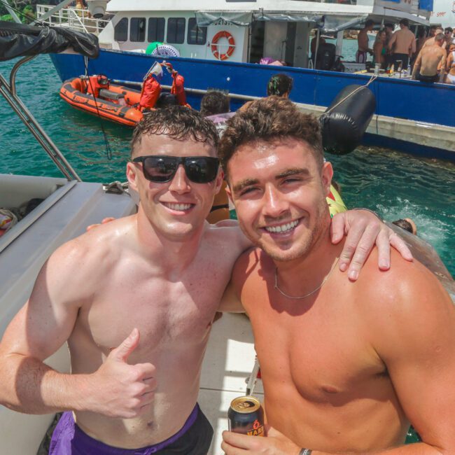 Two smiling men in swim trunks pose on a boat, one giving a thumbs up. There are people on nearby boats and in the water, with bright sunlight and clear blue water in the background.