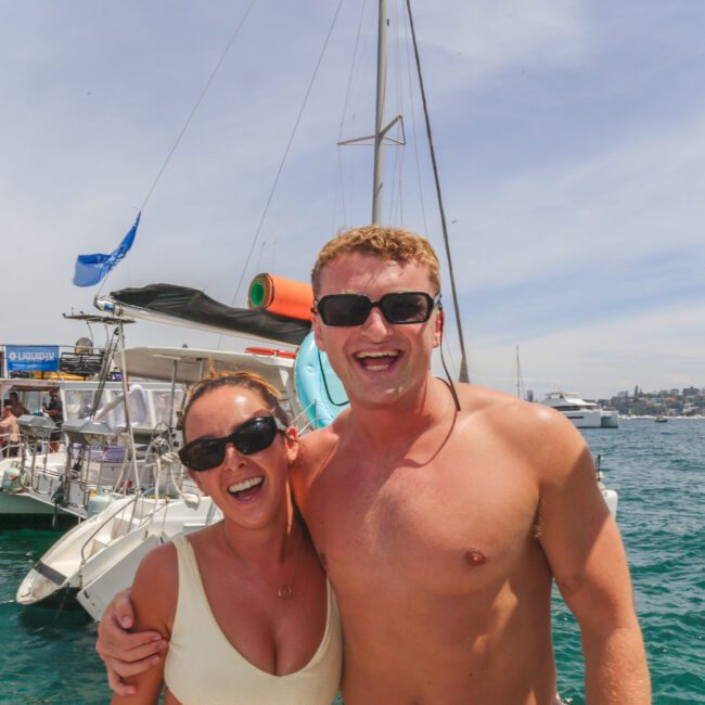 A smiling man and woman in sunglasses pose together on a boat, with other boats and people in the background on a sunny day at sea. The water is blue and the sky is partly cloudy.