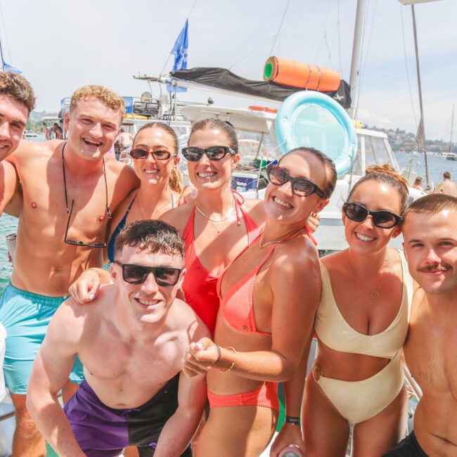 A group of young adults in swimsuits smile and pose together on a boat during a sunny day, with other people and boats visible on the water in the background.