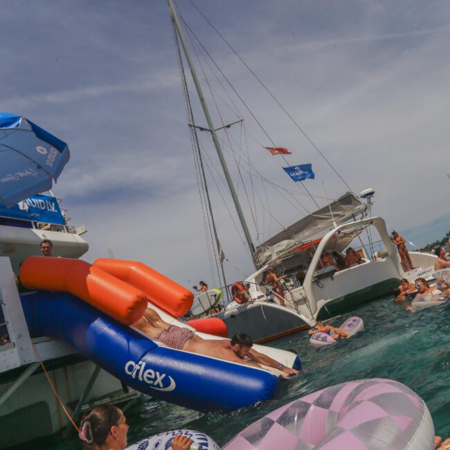 People enjoy a lively yacht party, floating on inflatables and relaxing in the water between boats under a sunny sky. Banners, umbrellas, and pool floats add to the festive atmosphere.