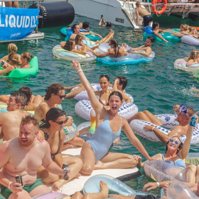 A group of people relax and have fun on colorful floaties in the water near anchored boats. Many are smiling, laughing, and raising their arms, enjoying a sunny day at a lively boat party.
