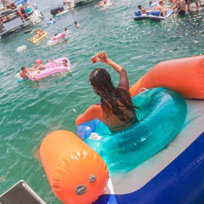 A woman sits on a colorful inflatable float, taking a selfie on a sunny day surrounded by people on various pool floats in the water near several boats. The scene is festive and lively.