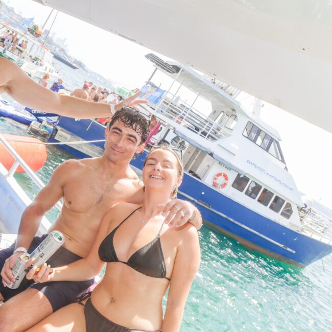 Three young adults in swimsuits smile on a boat; two are holding drinks. There’s a large blue-and-white boat and other people in the water and on boats in the sunny background.