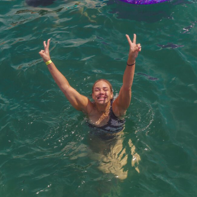 A smiling person in a swimsuit stands in clear green-blue water, raising both arms and flashing peace signs with their hands. There's a "Yacht Social Club" logo in the lower right corner.