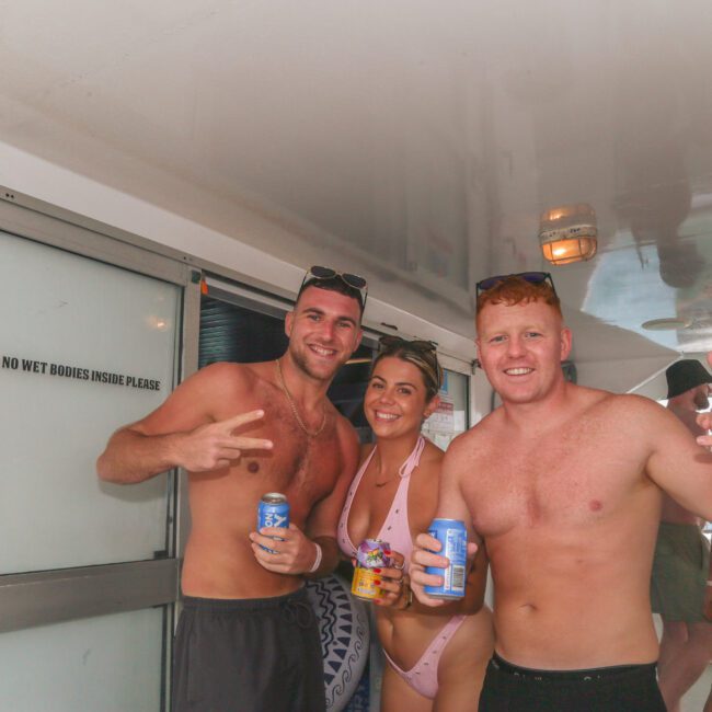 Three smiling people in swimwear stand on a boat holding drinks and snacks. Two men make hand gestures while a woman in the middle wears sunglasses. Other people are visible in the background. A “No wet bodies inside” sign is on the door.