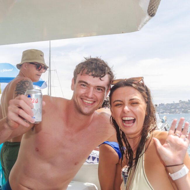 A man holding a can and a woman waving smile at the camera on a boat, with other people, umbrellas, and boats in the background on a sunny day by the water.