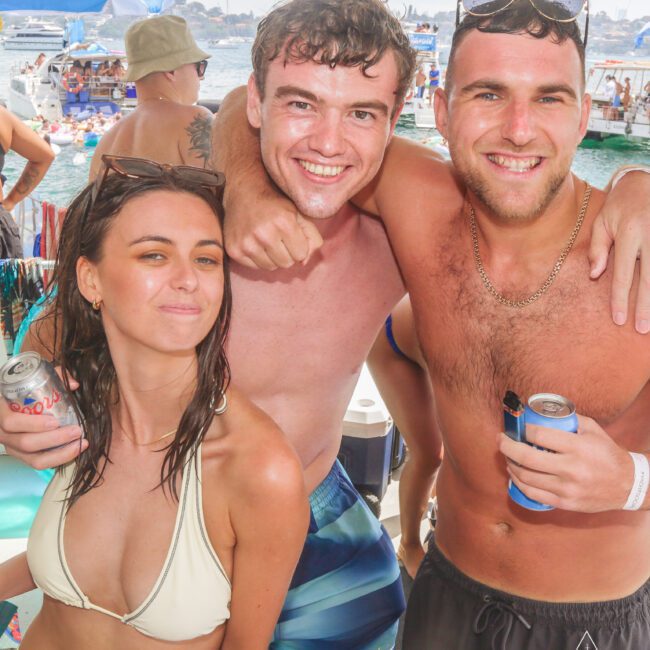 Three friends in swimwear smile and pose together on a boat, holding drinks. Other boats and people are visible in the sunny background, giving a lively and festive atmosphere.