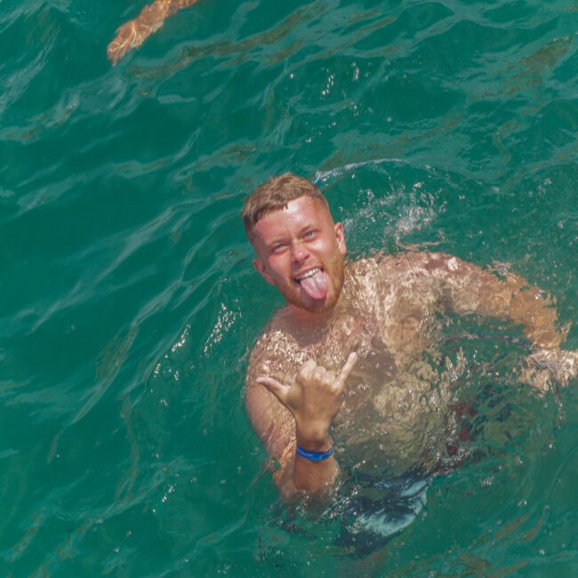 A man in swim trunks is in turquoise water, smiling, sticking out his tongue, and making a shaka hand sign. Another person's arm is visible at the top. "Yacht Social Club" is written in the corner.