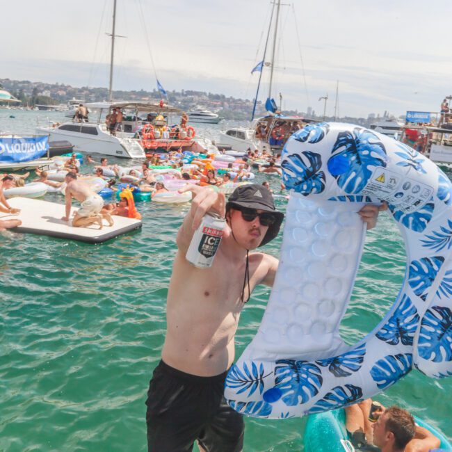 A young man wearing sunglasses and a hat holds a drink and a large inflatable in the shape of the letter "G" on a dock. Behind him, people relax on floating rafts and boats on the water during a sunny party.
