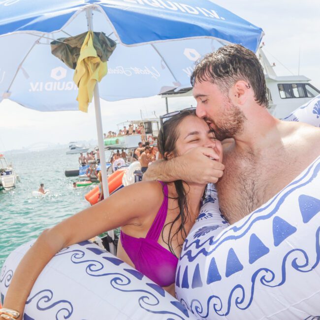 A smiling couple in swimsuits sit in inflatable floats, embracing and laughing, with boats and people in the background on a sunny day at a lively beach or boat party.