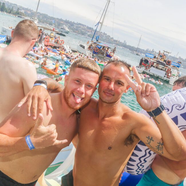 Two men in swim trunks smile and pose for the camera on a crowded boat, one making a peace sign and the other sticking out his tongue. People swim and float in the water nearby, with several boats in the background.
