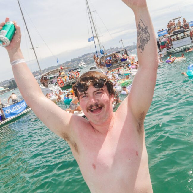 A man in red swim trunks, sunglasses, and a mustache raises his arms and smiles while standing in water. He holds a can, and boats with people are visible on the water in the background.