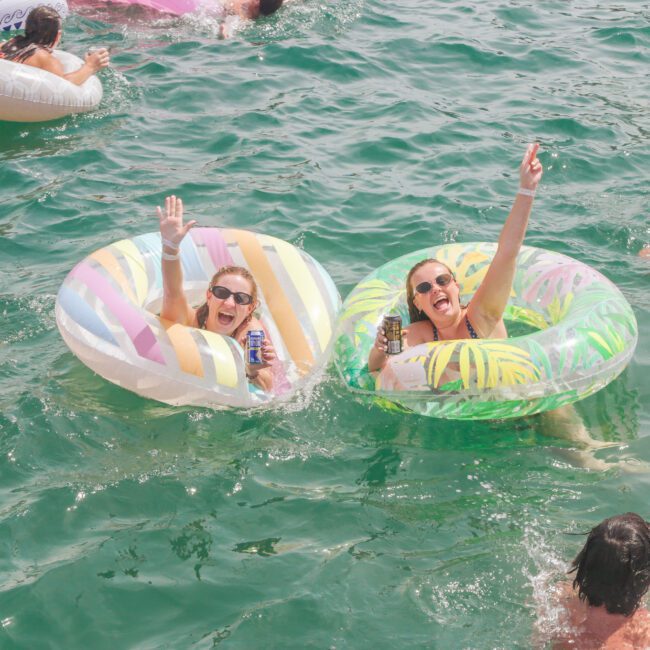 Two women in colorful inflatable tubes smile and wave while floating in green water. Other people swim and float around them, enjoying a sunny day. The scene is lively and playful.