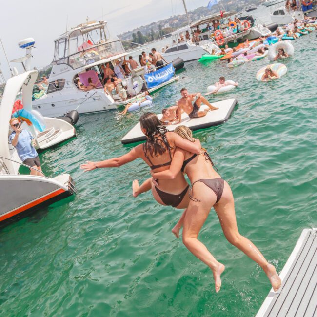 Two women in swimsuits jump into the water from a dock, surrounded by people swimming and lounging on boats and inflatables on a sunny day. The scene is lively and festive.