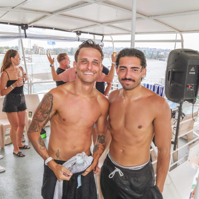 Two shirtless men in swim trunks smile at the camera on a boat during a party. Other people in swimwear are talking and enjoying themselves in the background, with water and buildings visible outside.