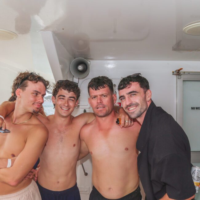 Four young men pose together, some shirtless, smiling on a boat with the sea in the background. Other people are visible nearby. A “No Wet Bodies Inside” sign and a “Yacht Social” logo are also in the image.