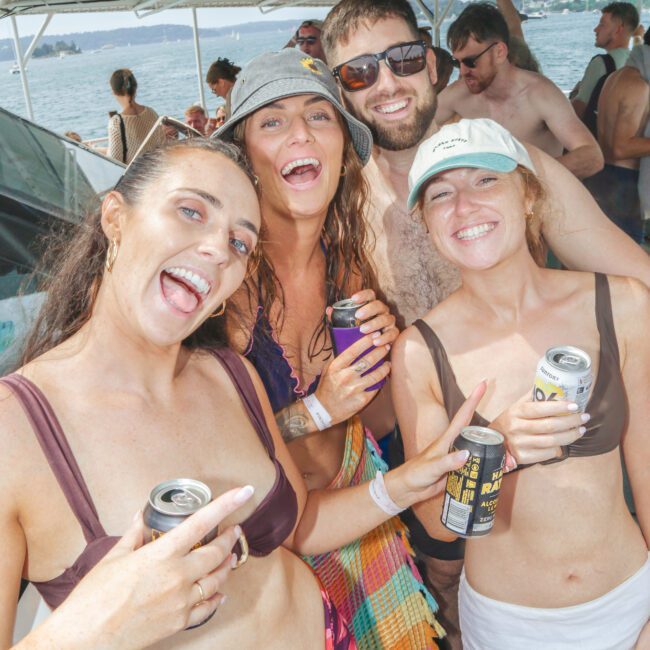 Four smiling people in swimwear pose together on a boat, each holding a canned drink. Water and other people are visible in the background, suggesting a lively, sunny boating event or party.