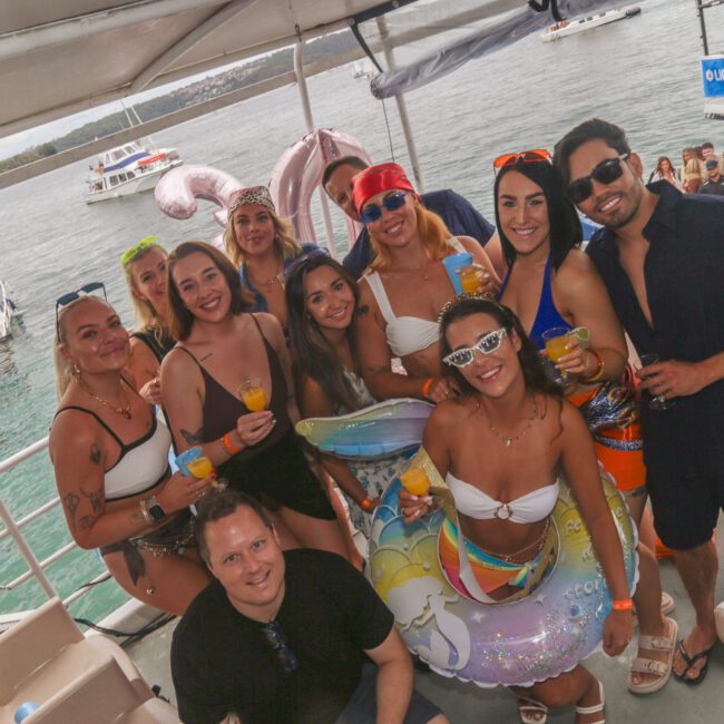 A group of people in swimsuits pose and smile on a boat, holding drinks. Some wear sunglasses and inflatable pool floats. Other boats and people can be seen on the water in the background. It’s a festive, summer atmosphere.