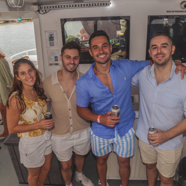 Four people stand close together on a boat, smiling at the camera and holding drinks. There are two men and a woman in light summer clothes, with water and other people visible in the background.