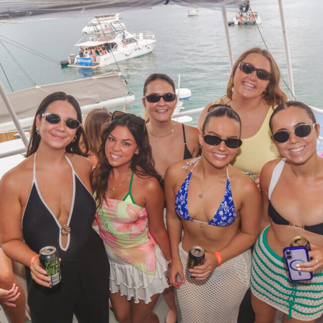 Six women in swimsuits and sunglasses smile at the camera on a boat. They are holding drinks, with water and more boats visible in the background. The mood is cheerful and relaxed, suggesting a fun day on the water.
