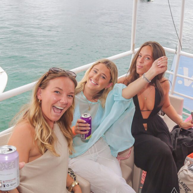 Three women smiling and enjoying drinks while sitting on a boat, with water and other boats visible in the background. They appear relaxed and cheerful, dressed in casual summer outfits.
