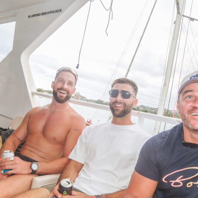 Four men sit and smile on a boat, enjoying drinks. One man is shirtless, the others wear t-shirts. The background shows a cloudy sky and water with distant buildings. The atmosphere is relaxed and cheerful.