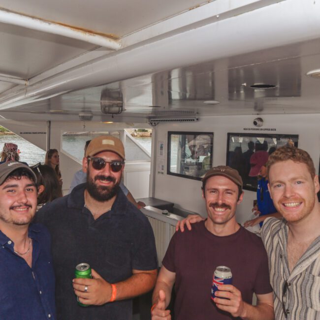Four smiling men pose together on a boat, each holding a drink, with other people socializing in the background. The scene is casual and lively, with water and shoreline visible outside.