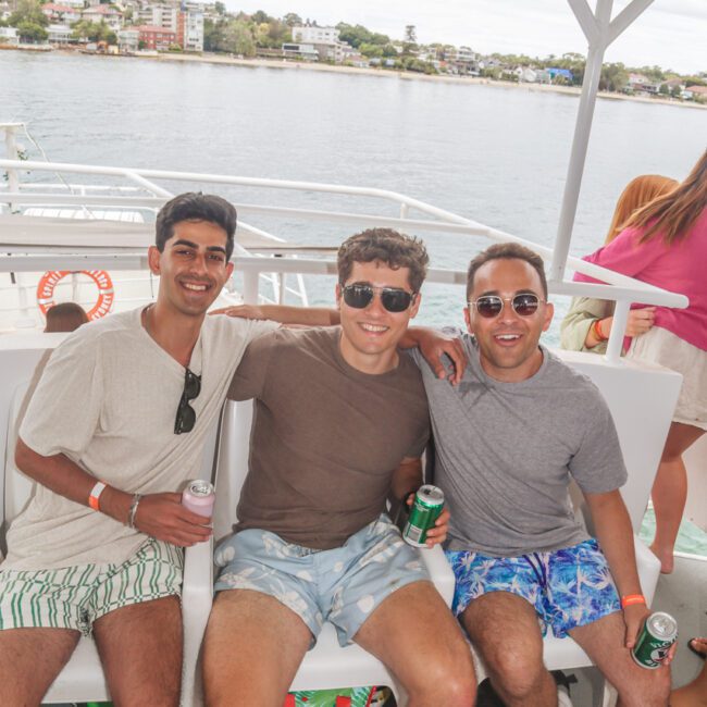 Three men sitting together on a boat bench, smiling and holding cans of drink. They are wearing summer clothes and sunglasses. The water, shoreline, and houses are visible in the background.