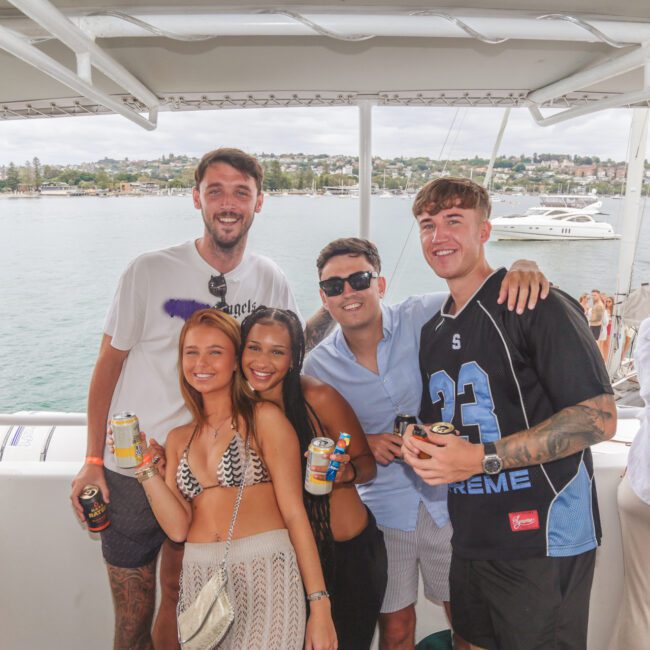 Five young adults, three women and two men, smile and pose together on a boat with drinks in hand. The background shows water, yachts, and a distant shoreline with buildings under a cloudy sky.