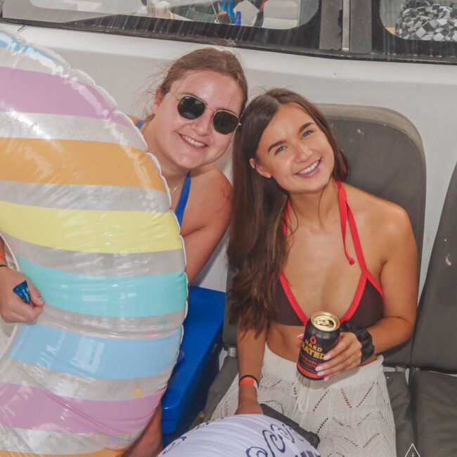 Two women sit on a boat, smiling at the camera. One wears sunglasses and holds a colorful inflatable ring; the other wears a red bikini top, white skirt, and holds a canned drink. Both look relaxed and happy.