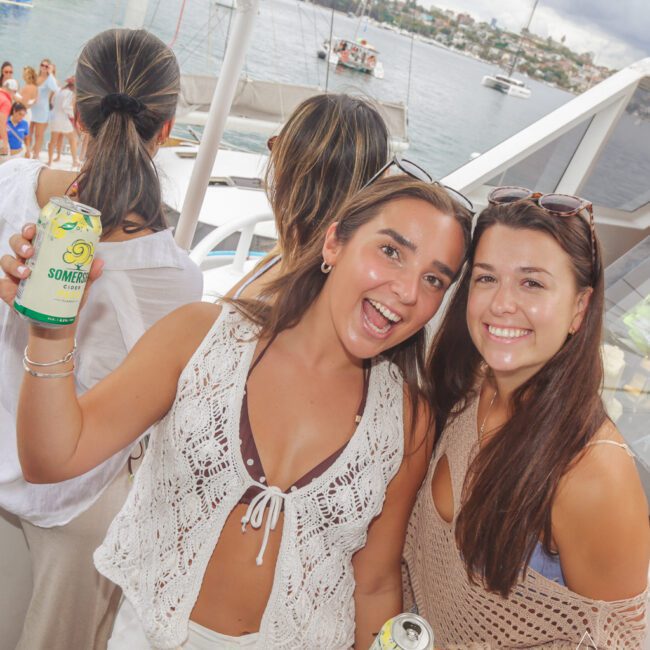 Two smiling women holding cans of Somersby cider pose on a boat, with water, yachts, and other people in the background, enjoying a lively outdoor event.