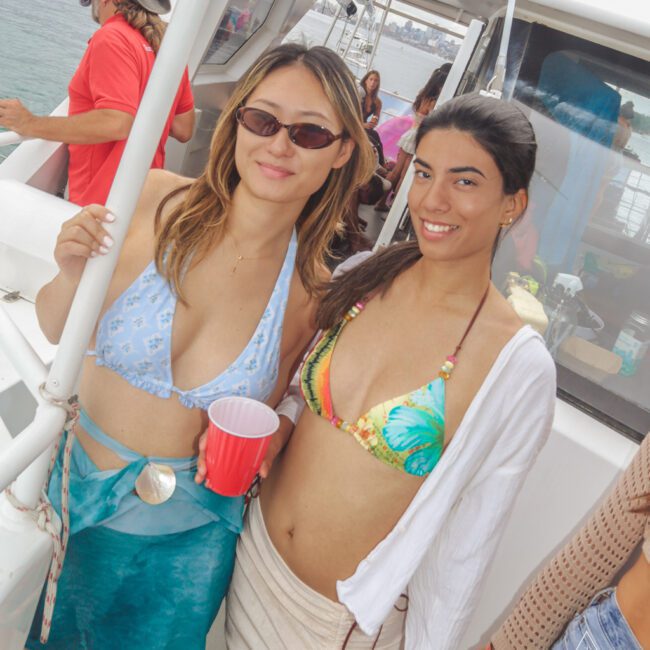 Two women in bikinis and sunglasses smile at the camera on a boat. One holds a red cup. Other people are visible in the background, with water and city views beyond the boat.