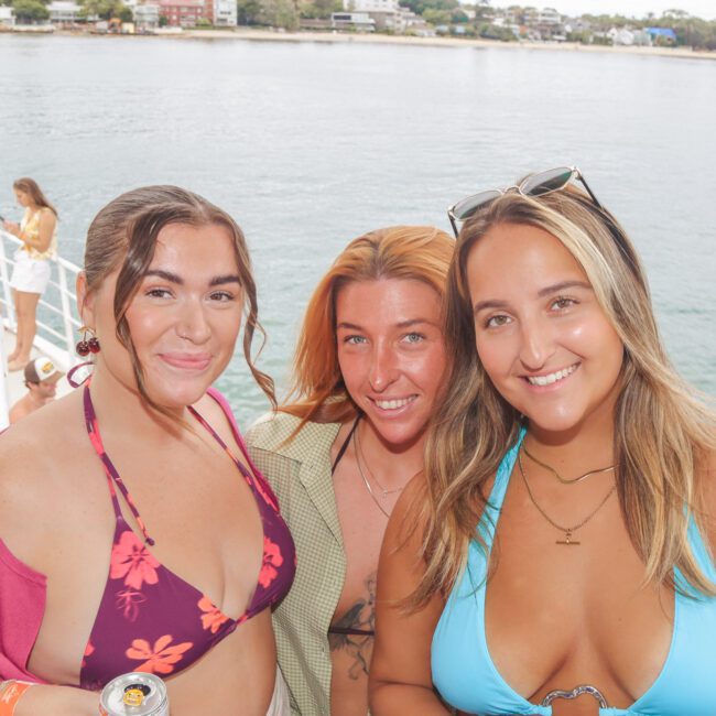 Three women in swimwear smile together on a boat with water and houses visible in the background. One holds a drink can, and the weather appears sunny and pleasant.